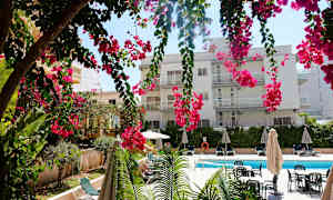 En bygning med flere balkonger bak hengende rosa blomster, et svømmebasseng og solsenger. (AI-generert tekst)