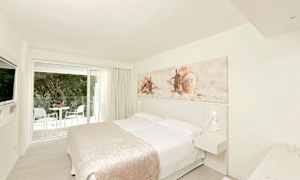 Et lyst hotellrom med en dobbeltseng, veggdekorasjon med skjellmotiv og et balkongområde utenfor glassdørene. (AI-generert tekst)