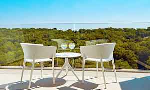 To hvite stoler ved et rundt bord med to glass på en veranda med utsikt over et skogkledd landskap. (AI-generert tekst)