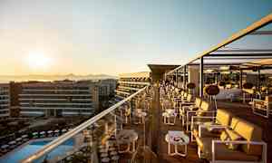 En takbar med sitteplasser og bord ved solnedgang, med utsikt over en basseng og bygninger. (AI-generert tekst)