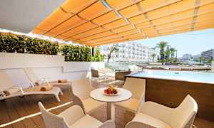 En utendørsterrasse med solsenger, bord og stoler. Det er et lite basseng og utsikt mot hotellbygninger. (AI-generert tekst)