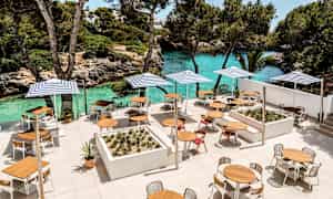 En terrasse med bord, stoler og parasoller ved en turkis vannstrand og grøntområder. (AI-generert tekst)