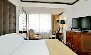 Et hotellrom med en seng med hvitt og beige sengeteppe, en lenestol og fotskammel ved vinduet, et skrivebord med stol og en TV plassert på en trekommode. (AI-generert tekst)