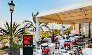 En utendørs restaurant med hvite duker og røde stoler under et tak, omgitt av palmer med utsikt over en marina. (AI-generert tekst)