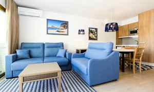 En stue med blå sofaer, et lavt bord, vegghengte malerier og et kjøkken med treverk i bakgrunnen. (AI-generert tekst)