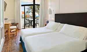 Et lyst hotellrom med to enkeltsenger, et skrivebord og en balkong med utsikt over havet og palmer. (AI-generert tekst)