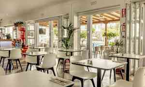 En restaurant med hvite stoler og bord, åpen mot en terrasse og omgivelser med grøntområder. (AI-generert tekst)