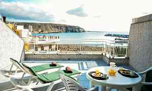 Terrasse med to solsenger og et bord med appelsinjuice og bakverk, havutsikt i bakgrunnen. (AI-generert tekst)