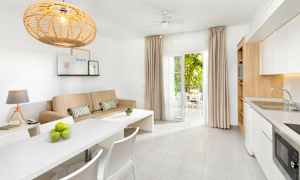 Et lys og moderne innredet rom med en beige sofa, hvitt spisebord og kjøkken, samt åpne dører til en møblert balkong. (AI-generert tekst)