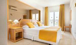 Et hotellrom med en dobbeltseng, hvite laken og gule puter og sengeteppe. Rommet har lyse tremøbler og vinduer med gardiner. (AI-generert tekst)