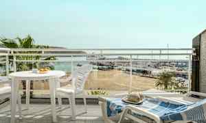 Utsikt fra en balkong med hvite utemøbler, med utsyn over en strand, en småbåthavn og havet i bakgrunnen. (AI-generert tekst)