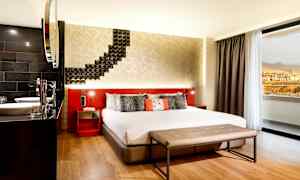 Et hotellrom med en stor seng, moderne interiør og en sittebenk foran sengen. Et vindu viser utsikten mot fjell og bygninger. (AI-generert tekst)