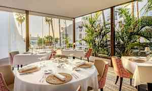 En restaurant med dekket bord, store vinduer og utsikt over palmer. (AI-generert tekst)
