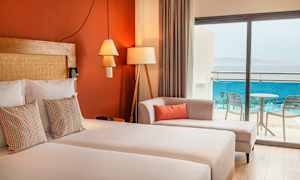 Et hotellrom med dobbeltseng, dekorative puter, en sofa og glassdører som fører til balkongen med utsikt over havet. (AI-generert tekst)