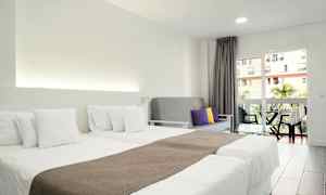 Et hotellrom med to enkeltsenger med hvitt sengetøy, en grå sofa med fargede puter og en balkong med utsikt. (AI-generert tekst)