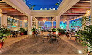 En utendørs servering med bord og stoler under en takkonstruksjon, omgitt av palmer og belysning. (AI-generert tekst)