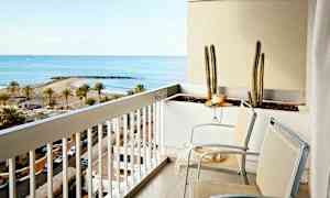 En balkong med to stoler og en blomsterkasse med kaktuser. Utsikten viser en strand med palmer og en bukt. (AI-generert tekst)