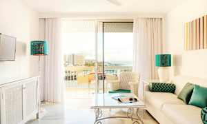 Lyst stue med en sofa, bord og stoler, samt glassdører som fører til en balkong med utsikt over havet. (AI-generert tekst)