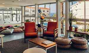 Et lyst rom med store vindusdører, to oransje stoler og kurvpaller foran et kaffebord. (AI-generert tekst)