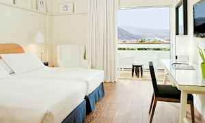 Et lyst og moderne hotellrom med to enkeltsenger, en hvit lenestol og et skrivebord med lampe. Et stort vindu fører ut til en balkong med utsikt over fjell og grønt. (AI-generert tekst)