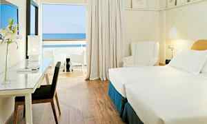 Et hotellrom med to enkeltsenger, skrivebord med stol, en hvit lenestol og utsikt mot havet fra en privat balkong. (AI-generert tekst)