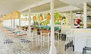 Et åpent uteområde med restaurantmøbler og lyse innredningsdetaljer, som hvite bord og stoler samt kurver. Dekorative lamper henger fra taket. (AI-generert tekst)