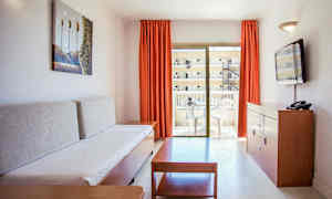 Stue i en ferieleilighet med en sofa, et salongbord, en kommode med en TV og en balkong med utemøbler. (AI-generert tekst)