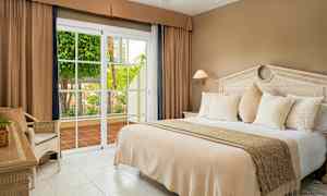 Et hotellrom med en stor seng, beige sengetøy, hvite puter og en glassdør som åpner mot en terrasse med grønne planter. (AI-generert tekst)