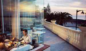 Par som spiser middag ved et bord nær glassvegg med utsikt over terrasse og hav. (AI-generert tekst)