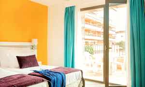 Et hotellrom med en seng, fargerike puter og tekstiler, samt en glassdør som fører til en balkong. (AI-generert tekst)