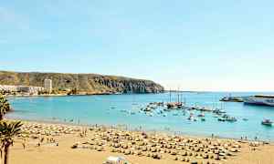 Strand med solsenger og parasoller ved en beskyttet havbukt med båter og bygninger synlige i bakgrunnen. (AI-generert tekst)