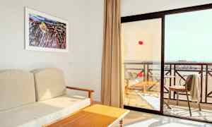 Utsikt fra et hotellrom med en beige sofa, et trebord og en balkong med utsikt over stranden. (AI-generert tekst)