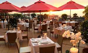 Utendørsrestaurant med røde parasoller, dekkede bord og solnedgang i bakgrunnen. (AI-generert tekst)