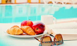 En tallerken med croissanter og epler står på et bord ved siden av en bok og et par solbriller. I bakgrunnen er et basseng synlig. (AI-generert tekst)