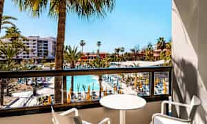 Utsikt fra en balkong over et hotellområde med basseng og solsenger, omgitt av palmer. (AI-generert tekst)