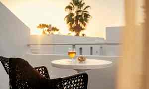 En balkong med et bord som har et glass med drikke og en skål med nøtter. I bakgrunnen sees en solnedgang og en palme. (AI-generert tekst)