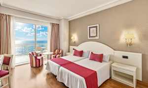 Et hotellrom med to senger, røde aksenter og havutsikt fra en balkong. (AI-generert tekst)