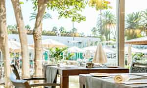 Bilde av et dekket bord ved et vindu med utsikt over trærne og parasoller på en solrik terrasse. (AI-generert tekst)