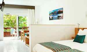 Lyst hotellrom med seng, bord og stoler samt utsikt mot en frodig balkong. (AI-generert tekst)