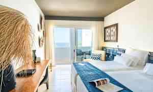 Et lyst hotellrom med to senger, en trebenk og en balkong med havutsikt. (AI-generert tekst)