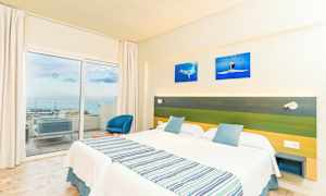 Hotellrom med to enkeltsenger, en grønn og blå sengegavl, samt en balkong med utsikt over havet. (AI-generert tekst)