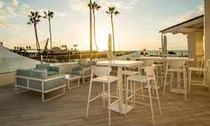 Utsikt over en møblert terrasse med bord og stoler ved solnedgangen, med palmer og havet i bakgrunnen. (AI-generert tekst)