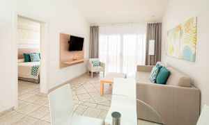 Bildet viser en lys og moderne stue med en sofa, TV samt spisebord og stoler. En dør fører til et soverom. (AI-generert tekst)