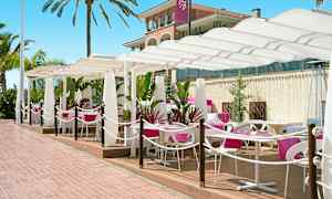 Utendørs servering med hvite parasoller og rosa duker på bordene. Palmer og en bygning i bakgrunnen. (AI-generert tekst)