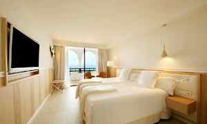 Et hotellrom med to senger, en veggmontert TV og en balkong med utsikt over havet. (AI-generert tekst)