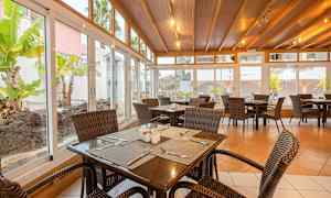 En spisesal med rottingstoler og trebord, omgitt av store glassvinduer med utsikt over en hage. (AI-generert tekst)