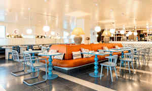 Et moderne innredet kafé med oransje skinnsofaer, blå bord og stoler samt dekorative lamper i taket. (AI-generert tekst)