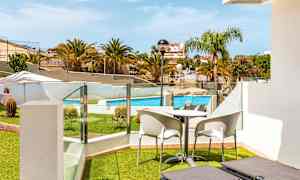 Utsikt fra en terrasse med solsenger, bord og stoler mot et basseng og omkringliggende grøntområder. (AI-generert tekst)