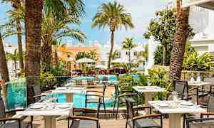 Bilde av en restaurantterrasse med utsikt over et basseng omgitt av palmer og lyse bygninger. (AI-generert tekst)