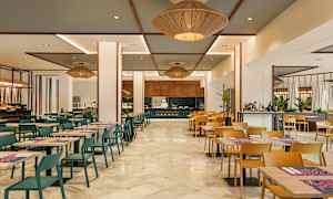 En spisesal med bord og stoler, der en kokk ses jobbe ved en matstasjon. (AI-generert tekst)